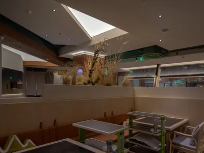 Modern restaurant interior with wooden tables, glass walls, and metal accents. Features an artificial plant centerpiece.