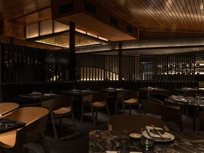 A modern restaurant interior featuring wooden tables and chairs, a wooden staircase, and a wooden ceiling.