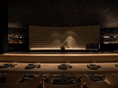 A modern restaurant interior with wooden tables and light-textured stone walls.