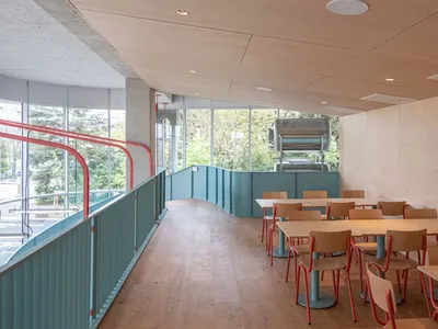 Industrial-themed restaurant interior with wooden tables and chairs, large windows and greenery outside.