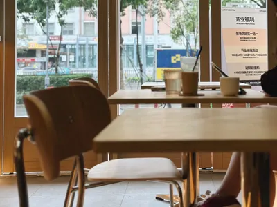 A cafe interior with modern wooden tables and chairs, featuring a window view of trees and buildings.