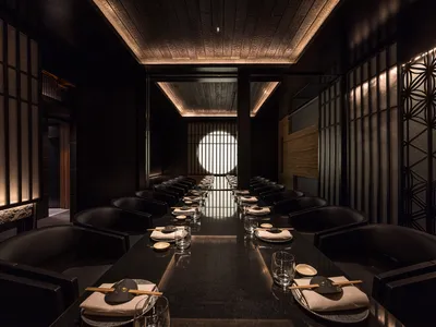 A modern restaurant interior with black furniture, featuring a long dining table set with white plates, chopsticks, and glass