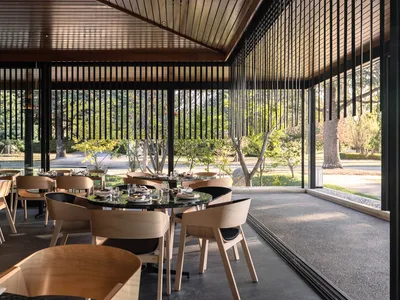 Modern restaurant interior with concrete flooring, wood furniture, and large windows overlooking greenery.