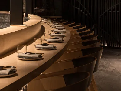 A modern restaurant interior featuring wooden tables and chairs arranged in a curved line, with soft lighting and a sleek des