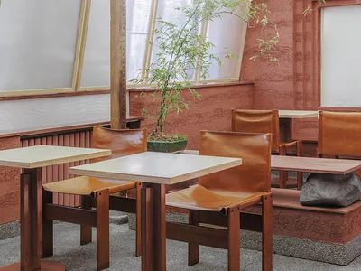 Modern restaurant interior featuring metal tables, leather chairs, cement flooring, and potted plants.