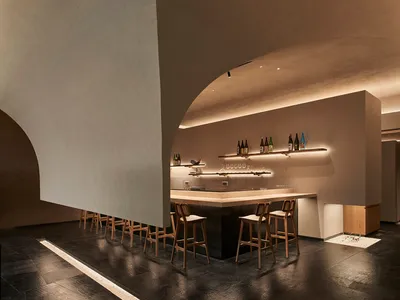 Modern, minimalist bar with concrete walls and wooden barstools, featuring a warm, inviting atmosphere.