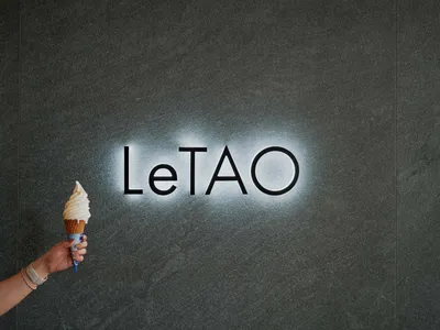 一隻手拿著冰淇淋甜筒，站在灰色大理石招牌前，背景有「LeTAO」燈光照亮。