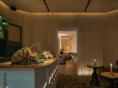 A modern restaurant interior featuring taupe curtains, wooden tables, and a central stone counter with a floral arrangement.