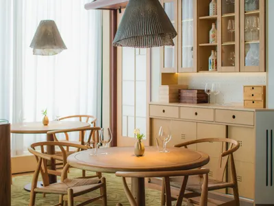 A modern restaurant interior with a wooden dining table and chairs, wooden counter, and hanging pendant lights.