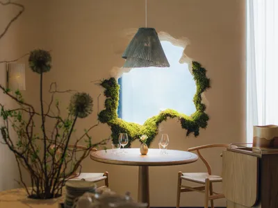 A modern restaurant interior with natural lighting, wooden tables, chairs, and a hanging pendant light surrounded by green mo