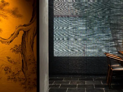 A minimalist restaurant interior featuring a wooden table, black cushions, and a bamboo shade covering a large window.