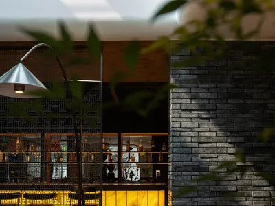 Modern industrial-style restaurant interior with a brick wall backdrop, round wooden dining table, and a bar or lounge area i