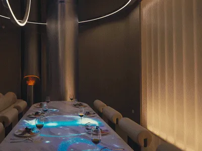 An empty industrial-style restaurant with a long dining table set for a meal and dim lighting.