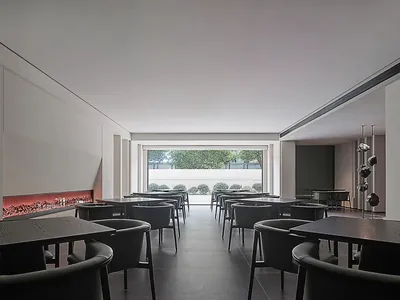 A sleek, minimalist restaurant interior featuring modern concrete dining tables and chairs arranged in rows, with a large flo