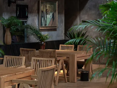 Industrial-style restaurant with concrete walls, wooden tables and chairs, metal railings, and potted plants.