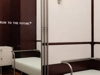 A minimalist hotel room featuring a metal frame bed with a light-colored mattress, placed on a wooden floor with a beige rug.