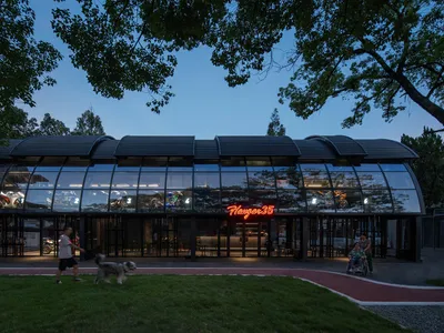 A modern restaurant exterior with large glass windows, illuminated signage, and a dog walking on the grass.