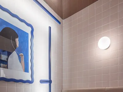 A minimalist restaurant interior featuring ceramic tiles, a bench, and a blue stool under a wall-mounted light.