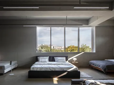 Modern minimalist bedroom with concrete walls, large windows overlooking greenery, and a king-sized bed with white linens.