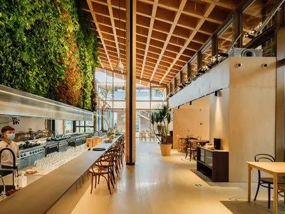 A modern restaurant interior featuring wooden tables and chairs, a living green wall, and large windows.