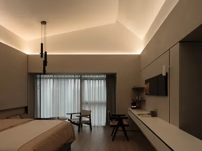 A modern minimalist bedroom with neutral-toned materials, featuring a bed, a desk, chairs, and a wall-mounted work area.