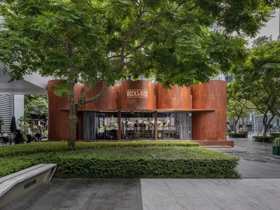A modern restaurant exterior featuring a metal corrugated facade under a lush tree canopy.