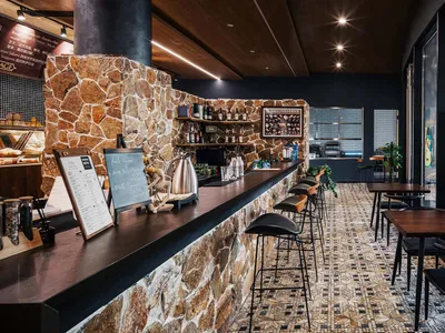 A modern bar area with stone walls, concrete flooring, and mosaic tile accents, featuring stools and a counter.