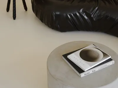 A minimalist black leather sofa in a living room setting, next to a vintage wooden stool and a concrete table with books.