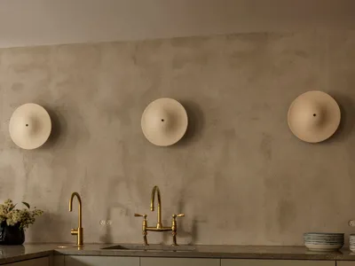 A modern, minimalist residential kitchen featuring concrete countertops and walls, with brass faucets and a stool.