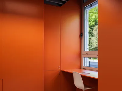 A modern home office featuring orange walls and a white chair in front of a window with greenery outside.
