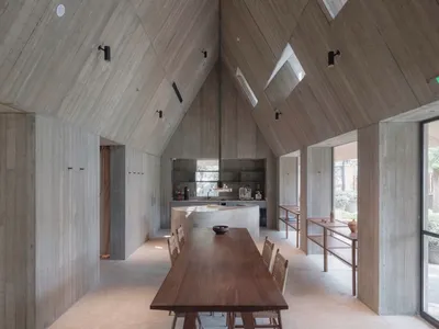 A minimalist residential kitchen featuring exposed concrete walls and ceiling, with a large wooden dining table and chairs.