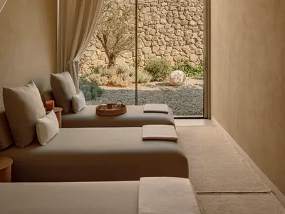 A minimalist bedroom with neutral tones, featuring twin beds, a window with curtains, and a view of a rocky garden.