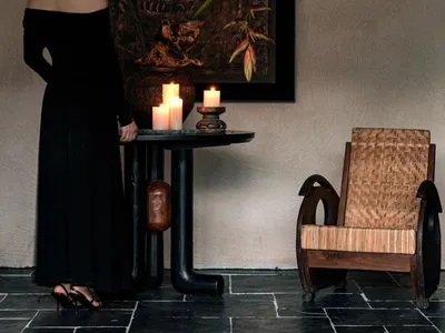 A woman in a black dress stands beside a table with candles and a textured vase in a living room setting.