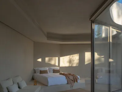 A minimalist bedroom featuring a concrete and glass platform bed, with white bedding and a window allowing sunlight to stream