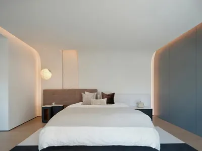 A modern minimalist bedroom with a king-sized bed, beige headboard, and white bedding. The room features a large window allow