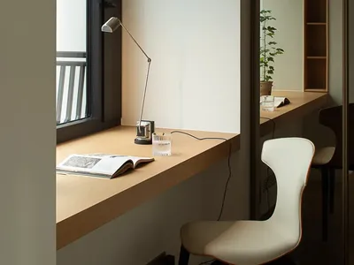 A minimalist home office featuring a wooden desk with a lamp and a book, white chair, and a window.
