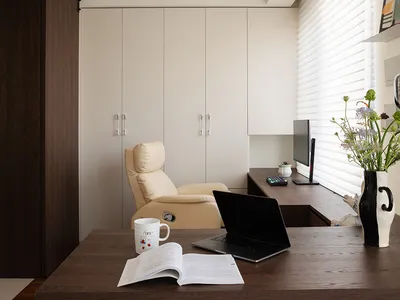 A modern minimalist home office with a wooden desk, beige leather chair, and a window with blinds.
