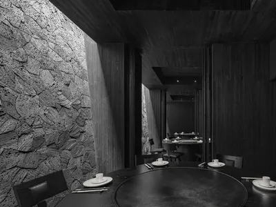 A modern restaurant interior with black tableware, chairs, and a stone wall, featuring a round table and minimalist decor.