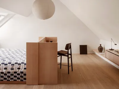 A minimalist bedroom featuring a wooden bed, a hanging light fixture, and a chair.