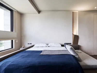 A minimalist bedroom featuring a blue bed with white sheets and pillows, set against minimalist white walls and a wooden floo