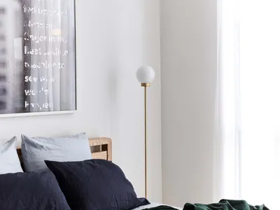 A minimalist bedroom with white walls, white flooring, and white blinds, featuring a bed with dark blue and green bedding, wh