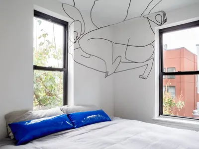 A minimalist bedroom with white walls, a bed with a blue pillow, and a wall mural of a black line drawing of a person