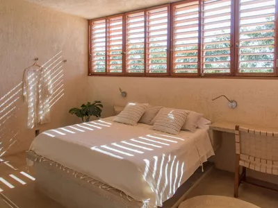 A minimalist bedroom featuring a concrete bed platform, white bedding, and closed shutters with sunlight filtering through.