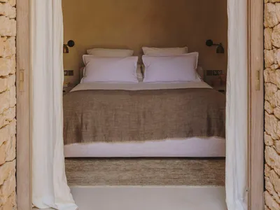 A minimalist bedroom with a bed covered in lavender linen sheets and a stone wall, enclosed by white curtains.