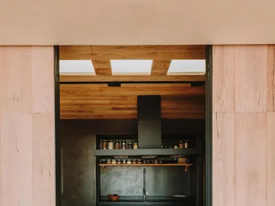 Modern kitchen with wooden ceiling and floor, featuring a black countertop, wooden stools, and a minimalist design.