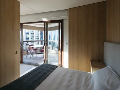 A modern minimalist bedroom featuring wooden walls and a white bed with gray accents, with a view of an outdoor seating area