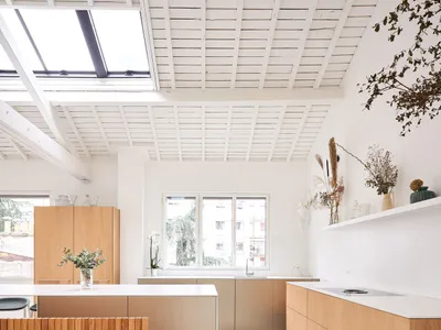 Modern kitchen features wooden cabinets and white flooring, with a minimalist design and large windows.