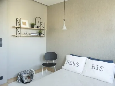 A minimalist bedroom with a bed, two pillows labeled 'HIS' and 'HERS', and a black chair with a shelf above it.