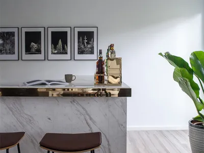 A minimalist bar studio featuring a marble counter with stools and a shelf of bottles and books.