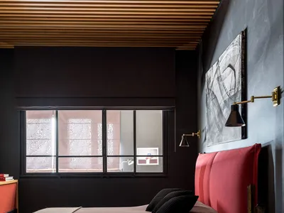 A modern minimalist bedroom featuring a red upholstered bed, dark walls, and a wooden ceiling.